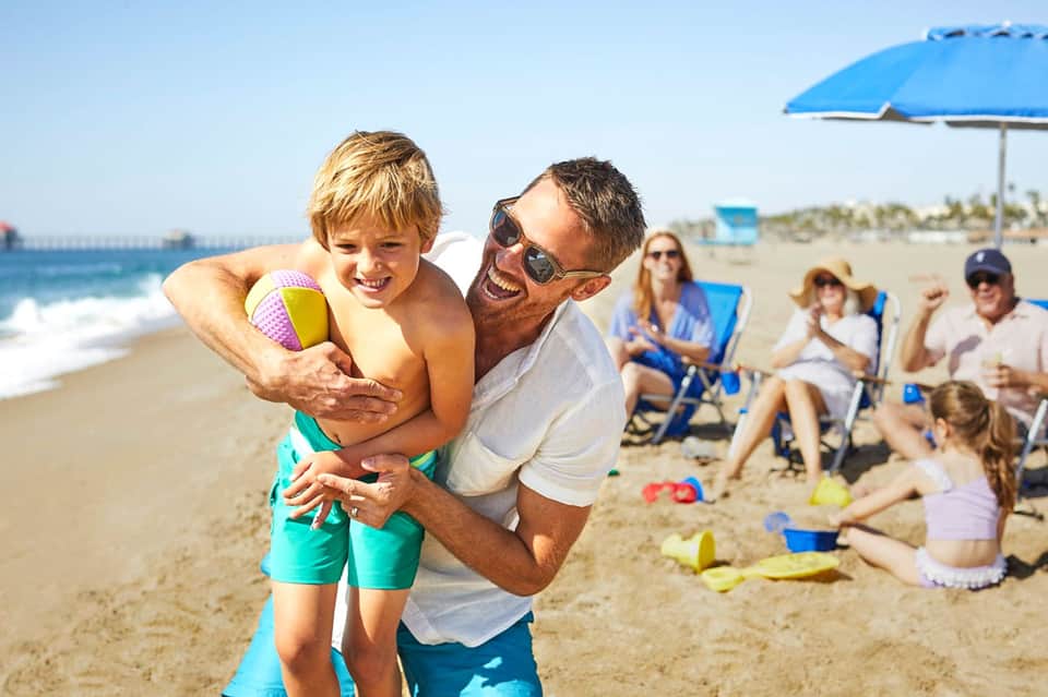 Father and young son playing together on sandy beach with families and umbrellas nearby