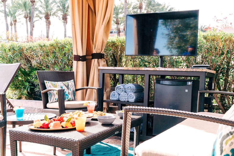 Outdoor patio with wicker furniture, orange curtains, TV mounted on wall, and tropical palm trees in background
