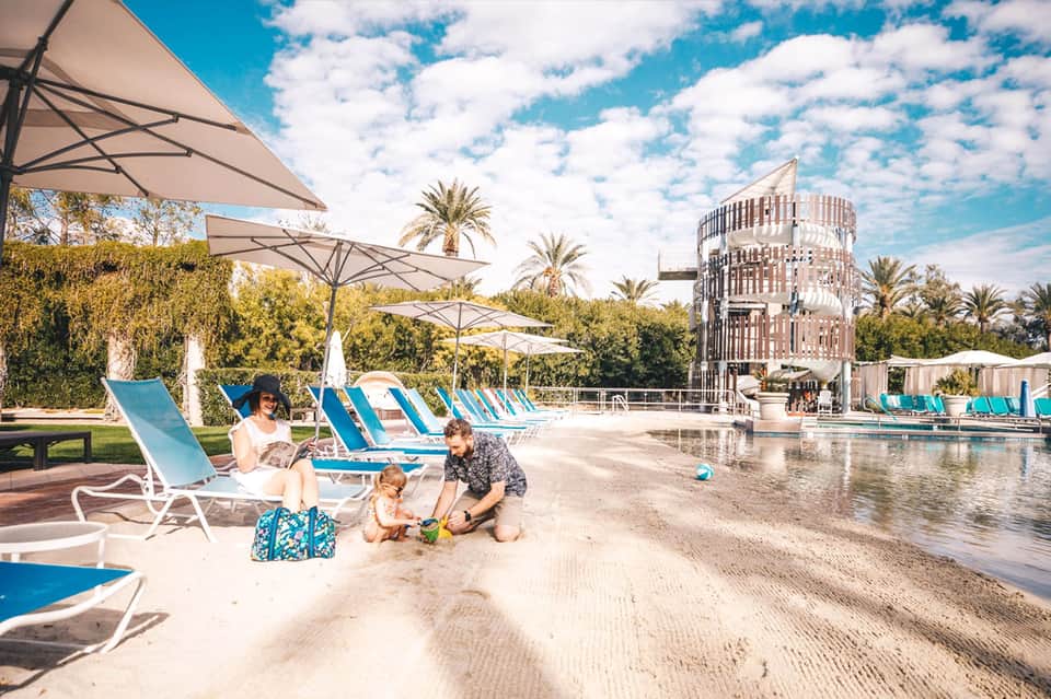 Family relaxing by a resort pool area with sandy beach, blue umbrellas, palm trees, and modern architecture