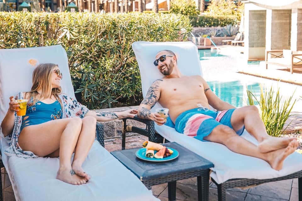 Couple relaxing on lounge chairs by poolside with tropical drinks and fruit plate on table