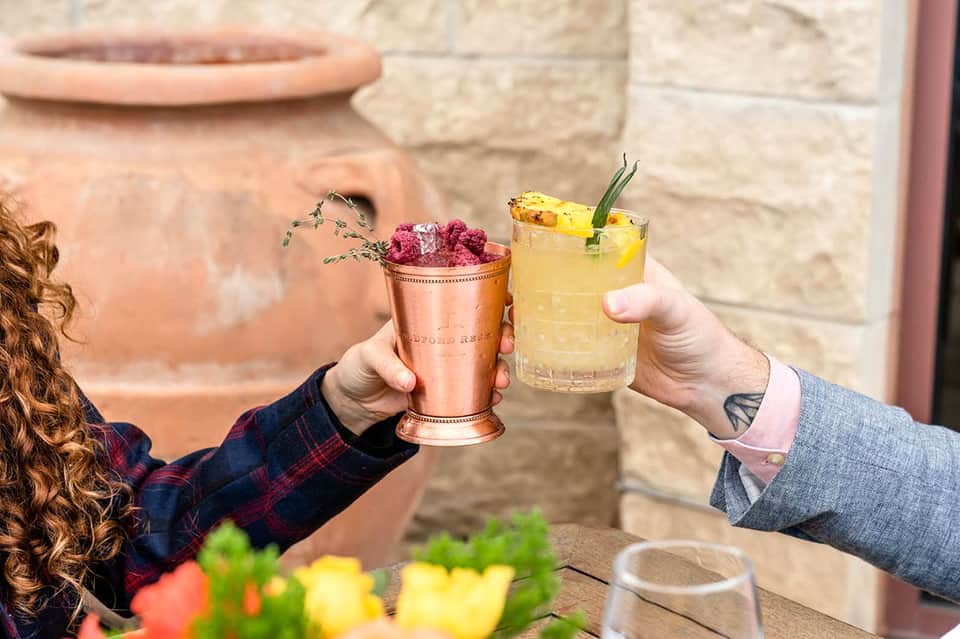 Two people toasting with colorful cocktails—a copper mule and a yellow drink.
