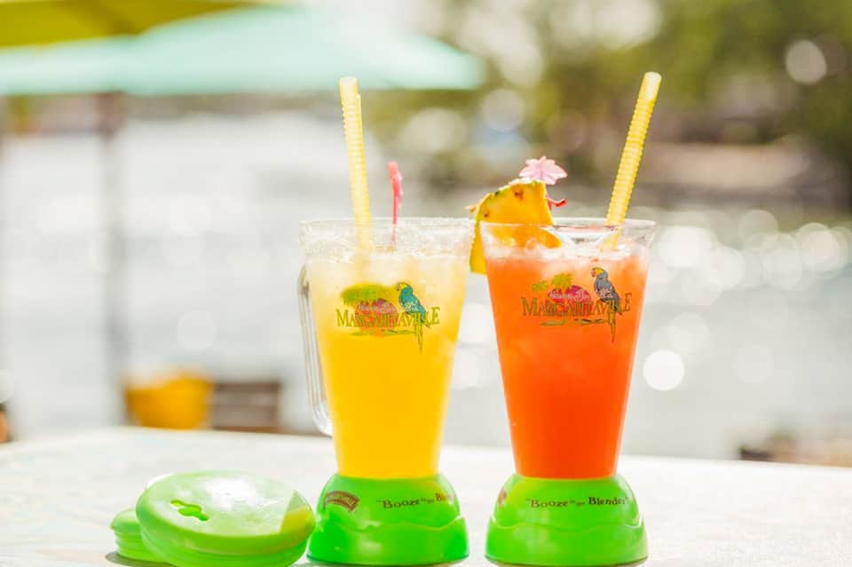Two colorful tropical cocktails with yellow and orange drinks, decorative straws, and green blender bases by the pool