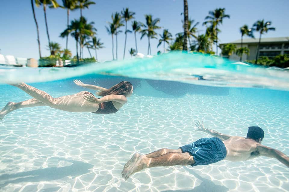 Two people snorkeling in crystal-clear turquoise shallow water with palm trees visible