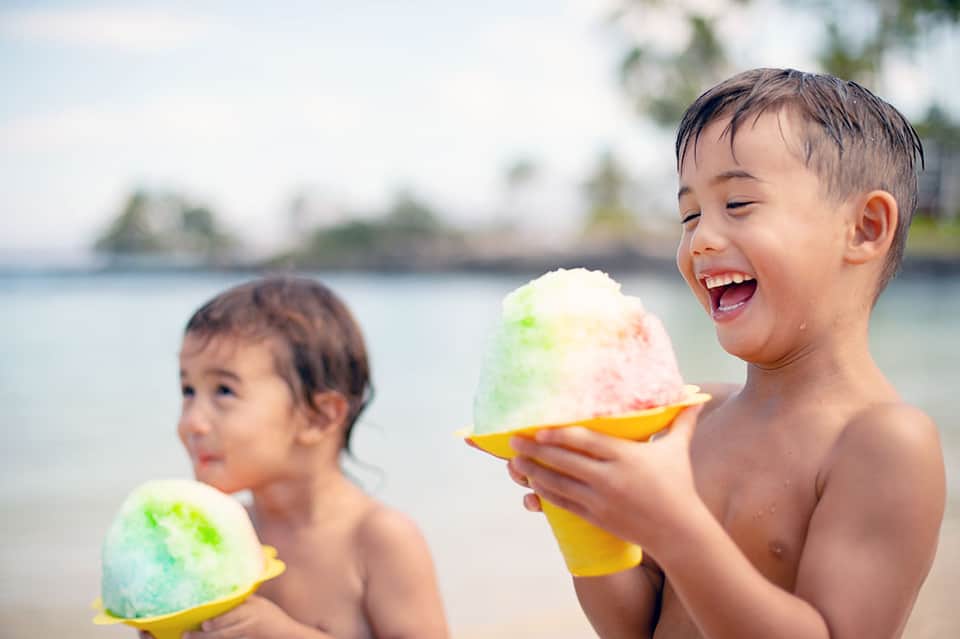 Two children enjoying colorful shaved ice treats on a tropical beach with palm trees