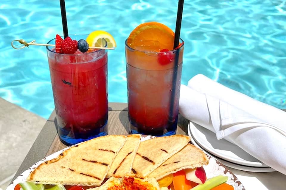 Two colorful tropical cocktails with fresh fruit garnishes beside pool with crackers and appetizers