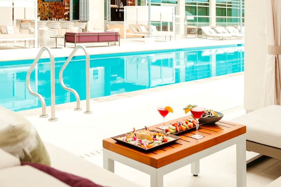 Modern indoor pool with glass enclosure, white lounge chairs, tropical plants, and appetizers on wooden table