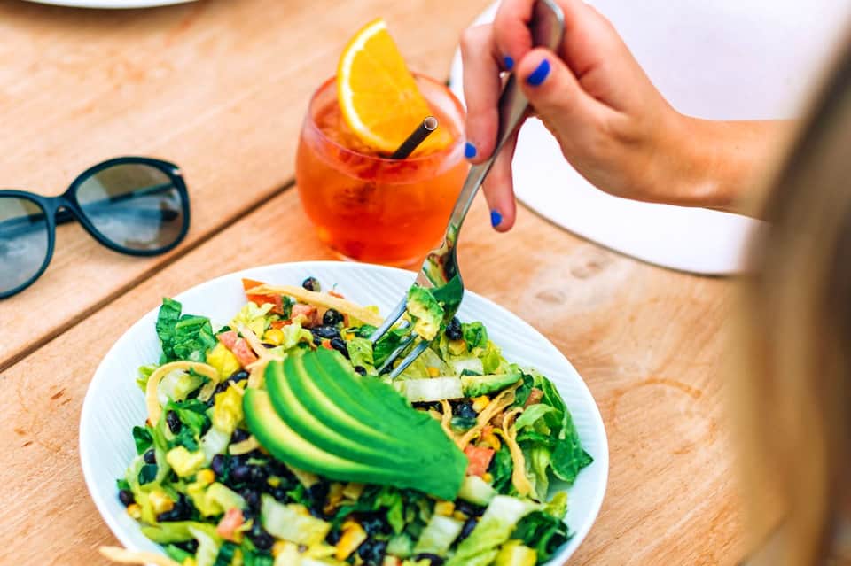 Woman dining outdoors with colorful salad and orange cocktail on wooden table with sunglasses nearby