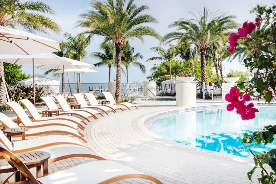 Luxury resort pool area with palm trees, white umbrellas, lounge chairs, and ocean view in background