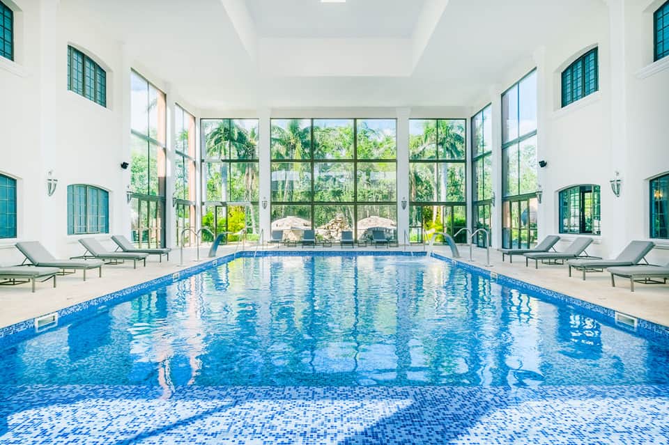 Indoor lap pool with high ceilings, large windows overlooking grounds, lounge chairs on sides