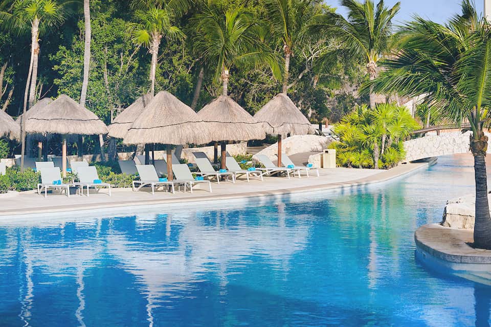 Resort pool with thatched umbrellas, white loungers, palm trees, and clear blue water