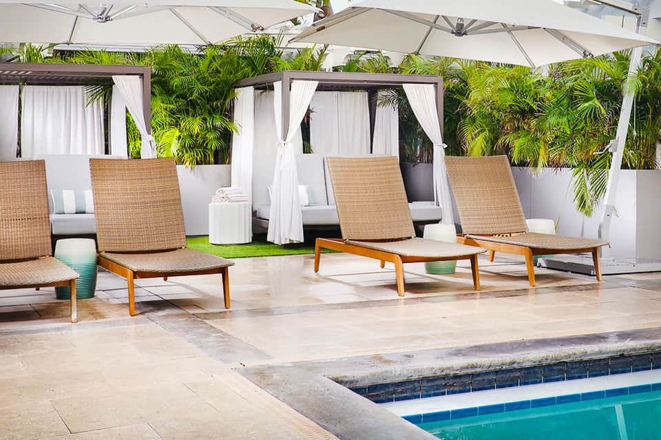 Outdoor lounge area with woven recliners under umbrellas, white curtains, and tropical greenery