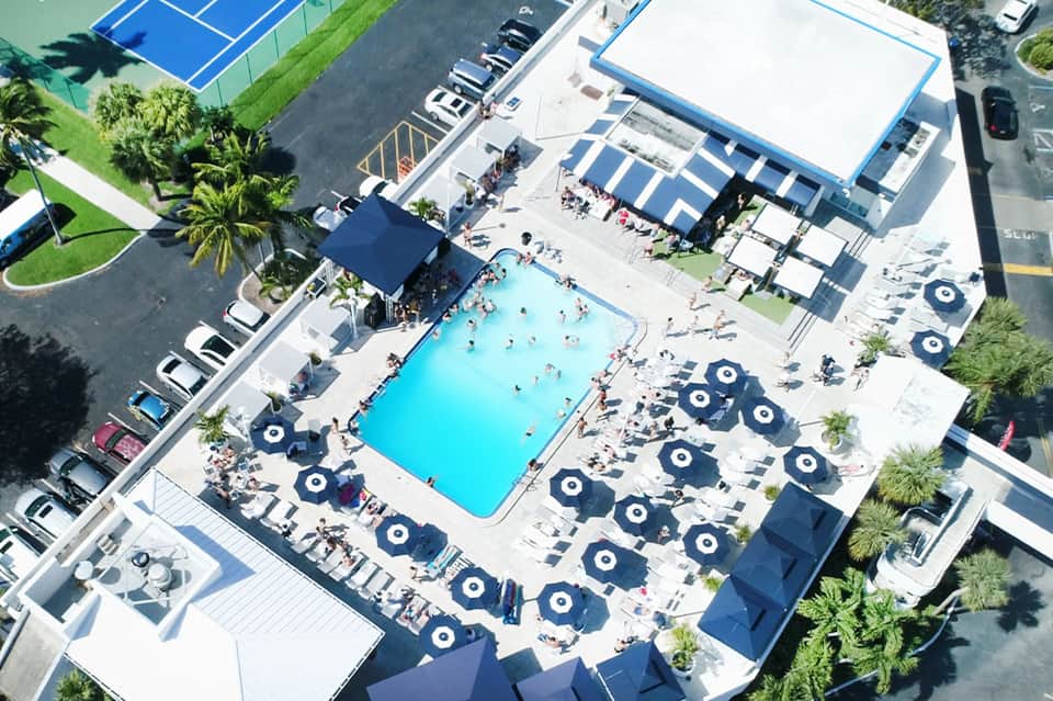 Aerial view of resort pool with bright blue water, umbrellas, and surrounding palm trees and facilities