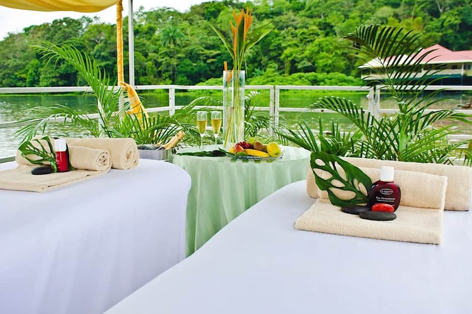 Spa treatment tables by waterside with tropical plants, towels, and wellness products on white tablecloths