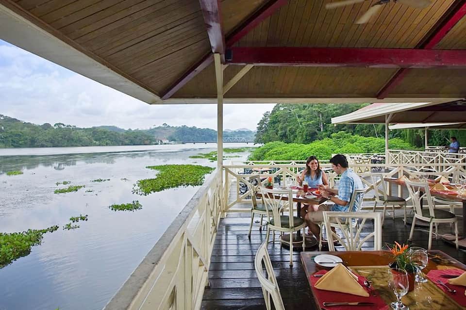 Waterfront dining deck with guests overlooking a river lined with tropical vegetation and mangrove trees