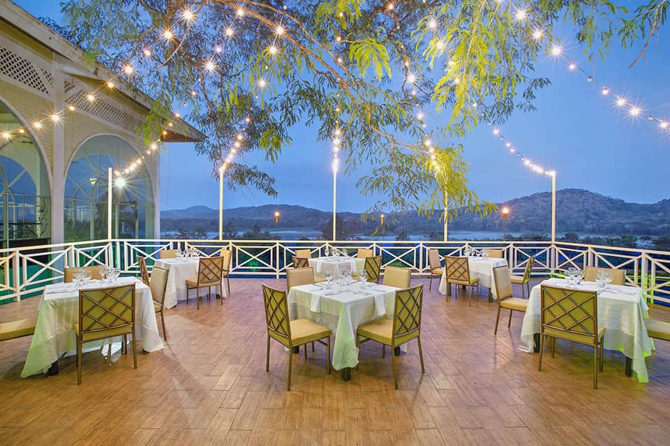 Elegant lakeside restaurant terrace at dusk with white tablecloths, green chairs, and mountain views