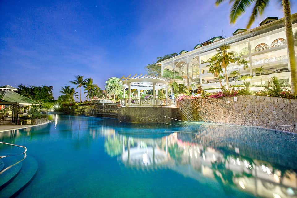 Illuminated resort pool at night with white architecture and palm trees reflected in water