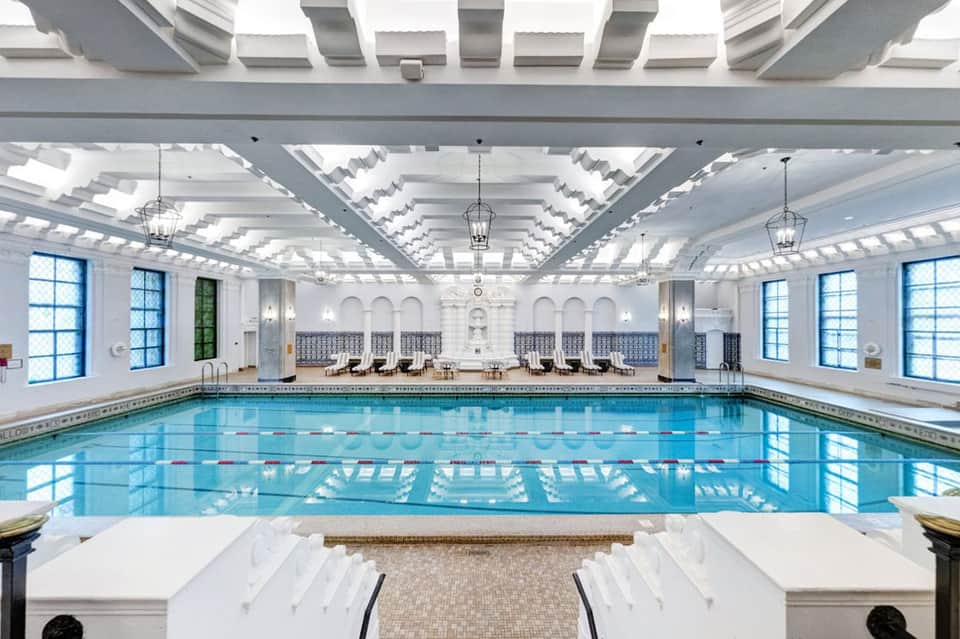 Indoor lap pool with lane markings, skylights, white columns, and lounge seating areas