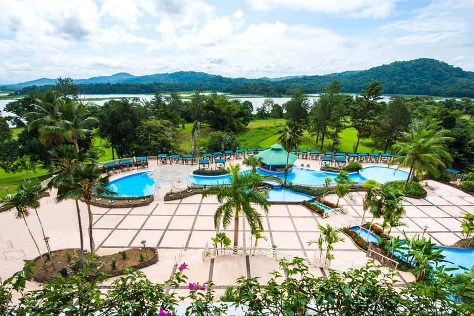 Aerial view of tropical resort with blue pools, palm trees, and river landscape with mountains