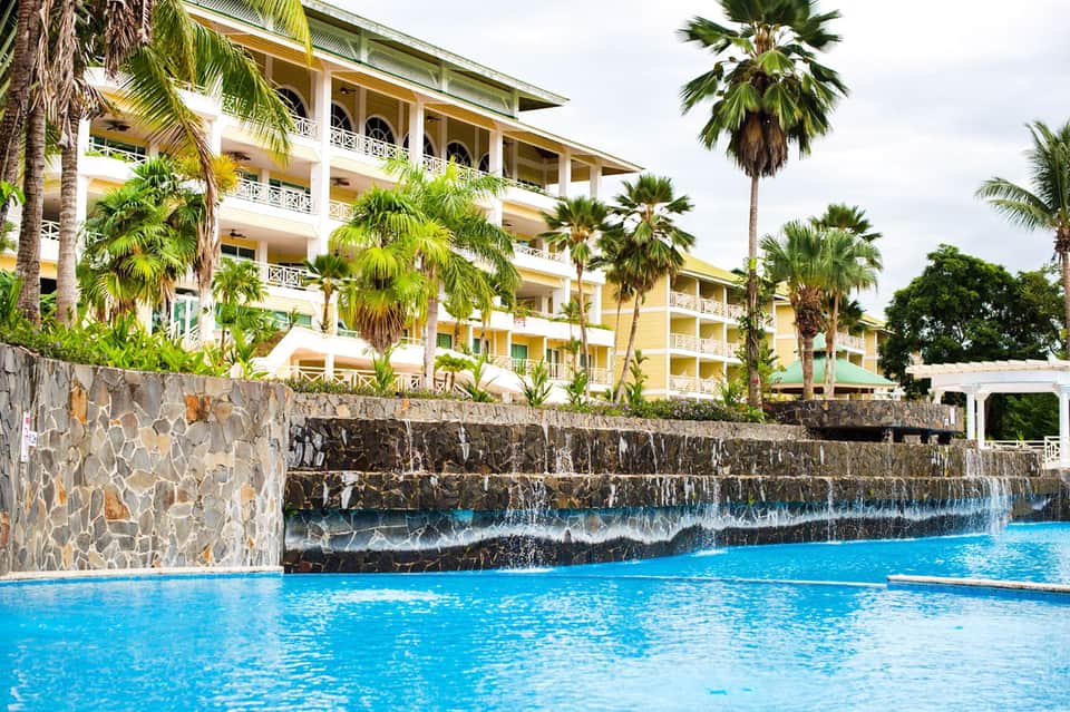 Tropical resort building with yellow exterior, palm trees, and blue swimming pool