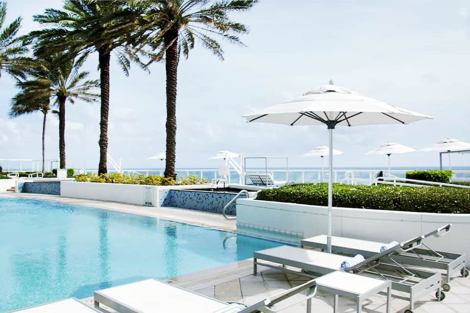 Beachfront pool with white umbrellas, palm trees, and ocean view