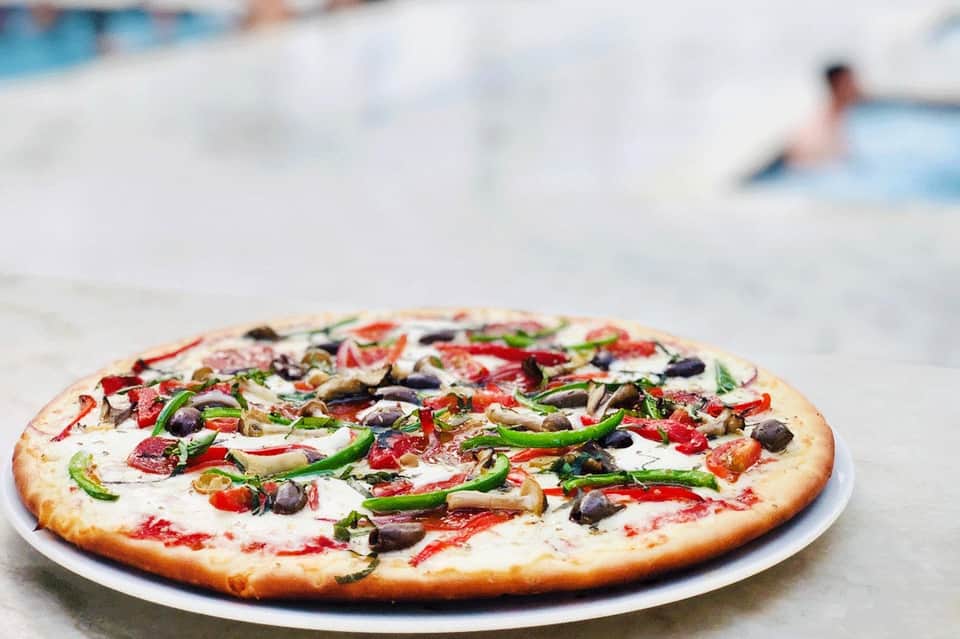 Colorful vegetable pizza with black olives and green peppers on white plate near beach