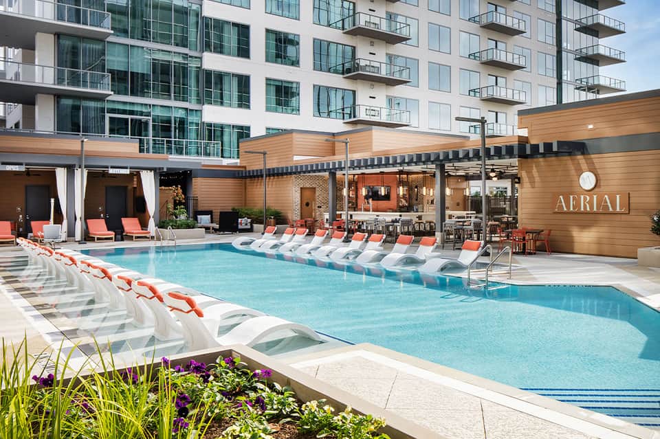 Modern hotel pool deck with loungers, striped umbrellas, wooden pergolas, and contemporary building with balconies