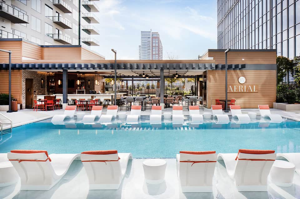 Modern hotel pool deck with loungers, wooden pavilion bar labeled Aerial, and city buildings