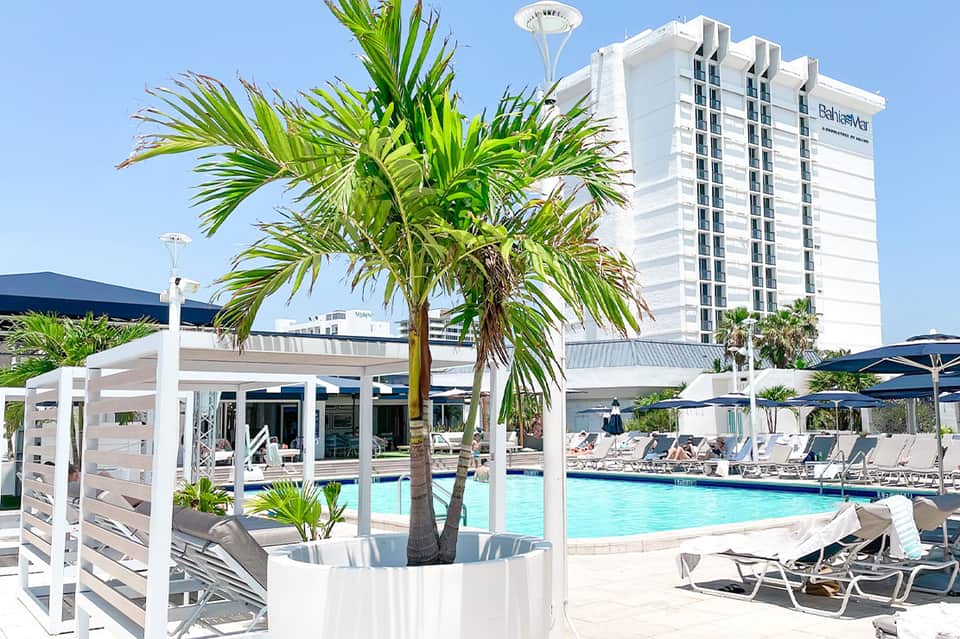 Modern resort pool area with white cabanas, palm trees, and high-rise hotel building