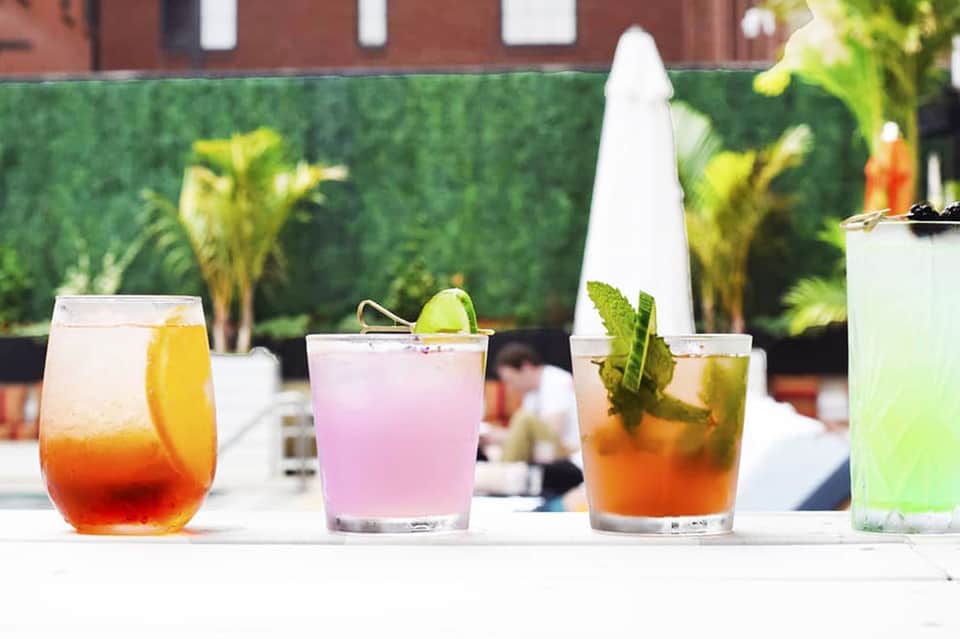 Four colorful cocktails displayed on white bar counter with green hedge background