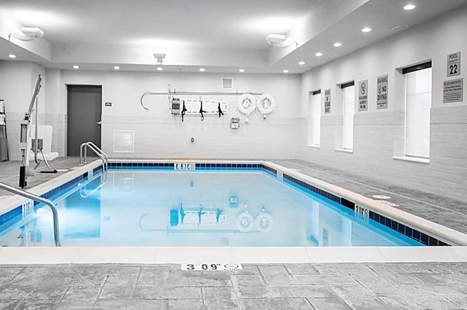 Indoor heated pool facility with blue water, white tile walls, windows, and pool safety equipment