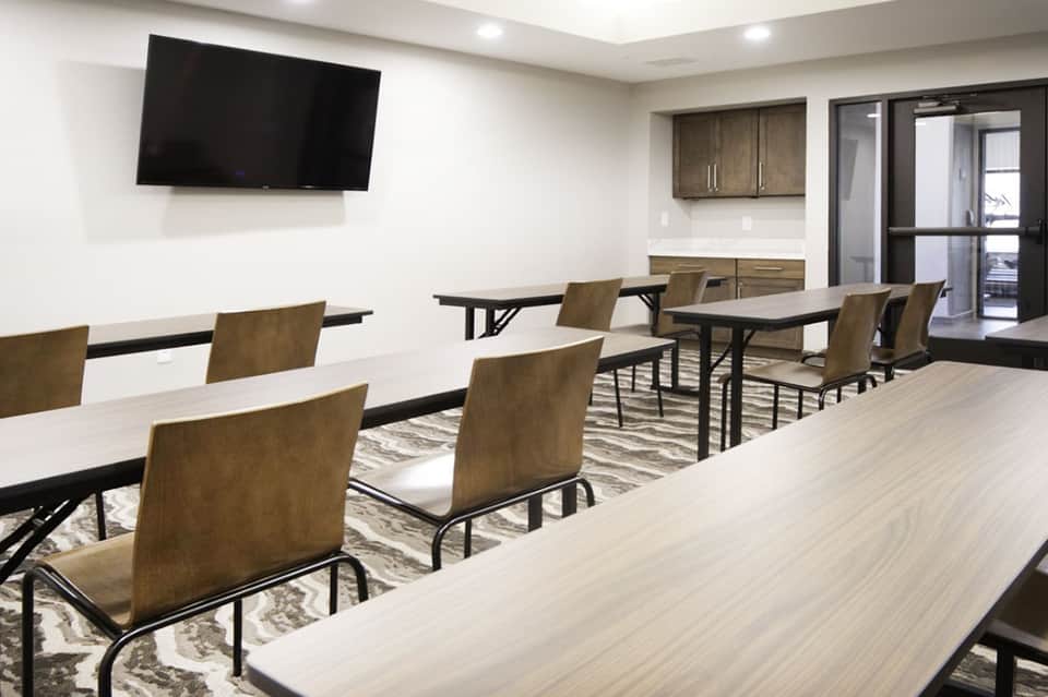 Modern meeting room with long tables, tan chairs, wall-mounted TV, and kitchenette