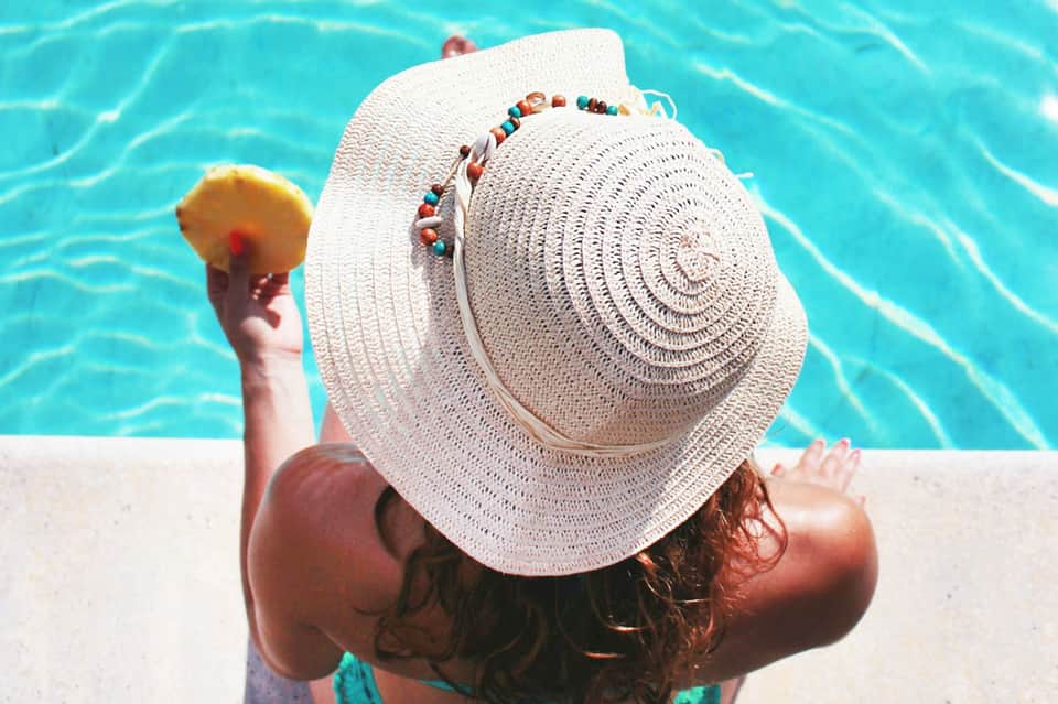 Person wearing white sun hat by turquoise pool water holding tropical drink