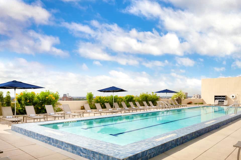 Modern rooftop lap pool with blue umbrellas, lounge chairs, and city skyline view