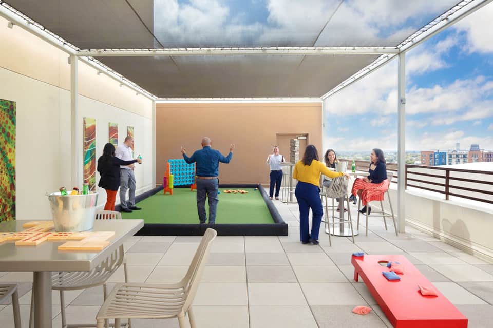 Rooftop lounge area with cornhole game, dining tables, and panoramic city views