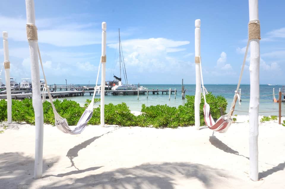 White hammocks strung between poles on sandy beach with sailboat and dock