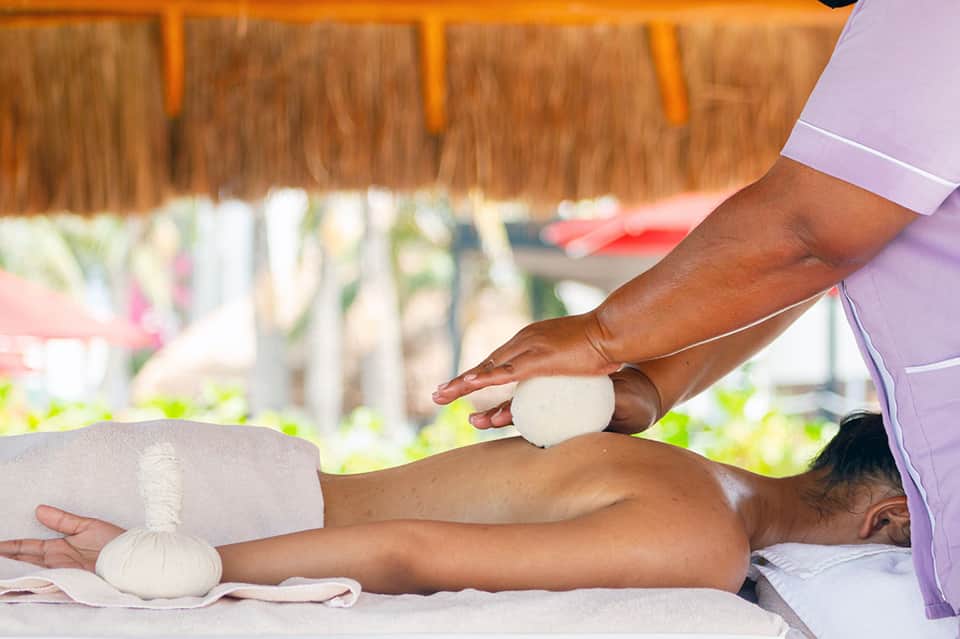 Spa therapist performing herbal massage treatment on client's back near beach