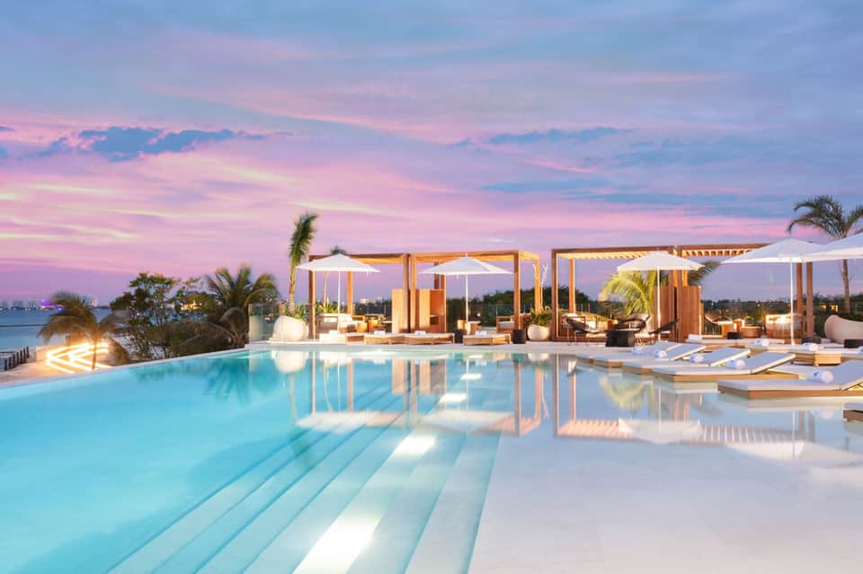 Infinity pool with wooden cabanas and lounge chairs at sunset overlooking water