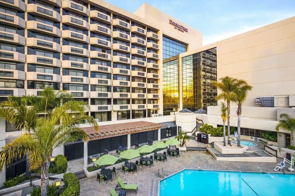 Hotel exterior with multi-story building, courtyard pool, green umbrellas, palm trees, and patio seating