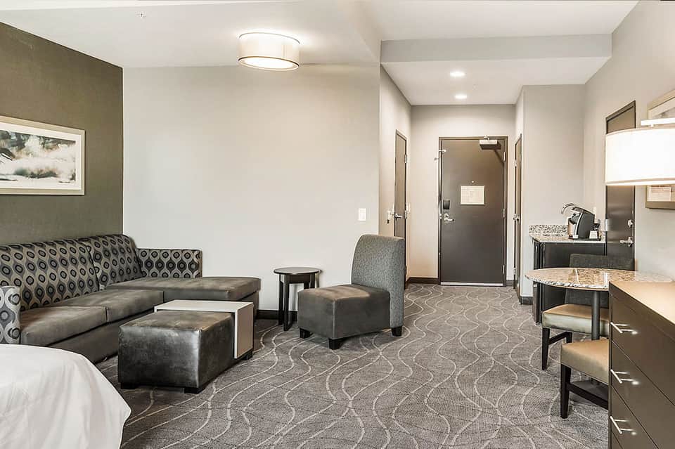 Hotel suite living room with patterned sofa, ottomans, kitchenette, and bathroom entrance