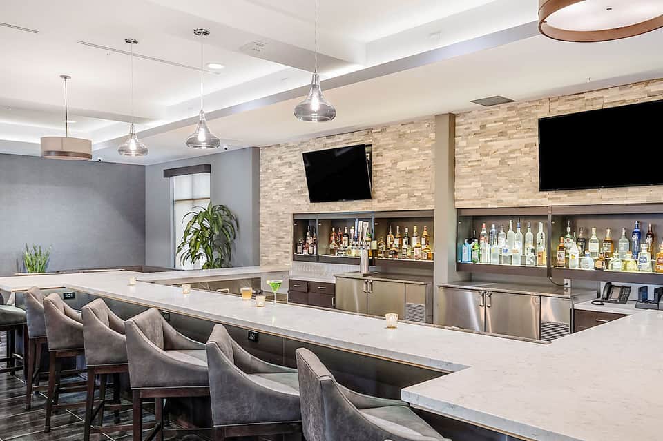 Modern hotel bar with brick accent wall, white countertop, pendant lighting, and illuminated liquor shelves