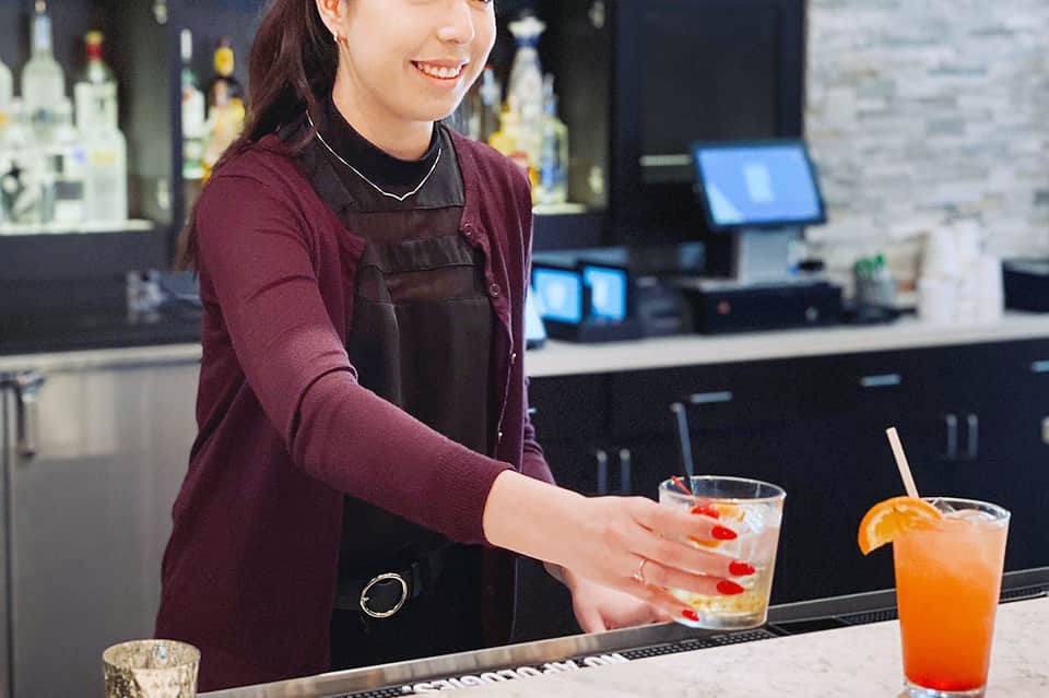 Female bartender in maroon shirt preparing cocktail at modern bar with bottles in background