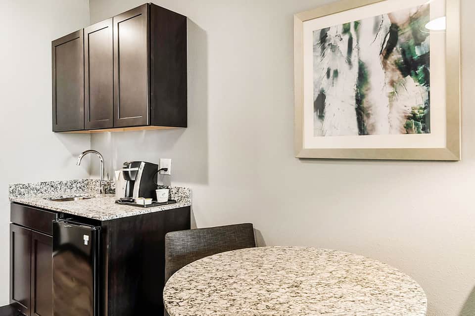 Modern hotel kitchenette with black cabinets, granite countertop, coffee maker, and round dining table