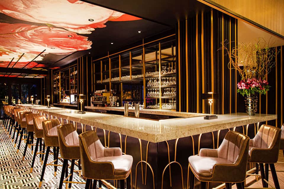 Elegant hotel bar with long counter, rose gold chairs, modern pendant lighting, and upscale décor