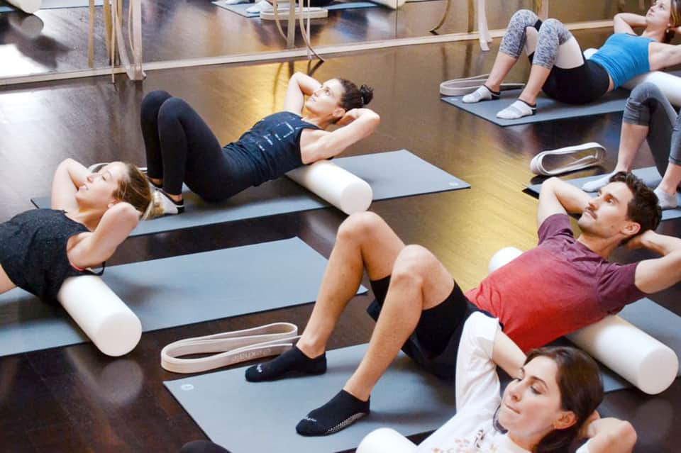 Group fitness class performing core exercises on yoga mats in a studio with mirrors