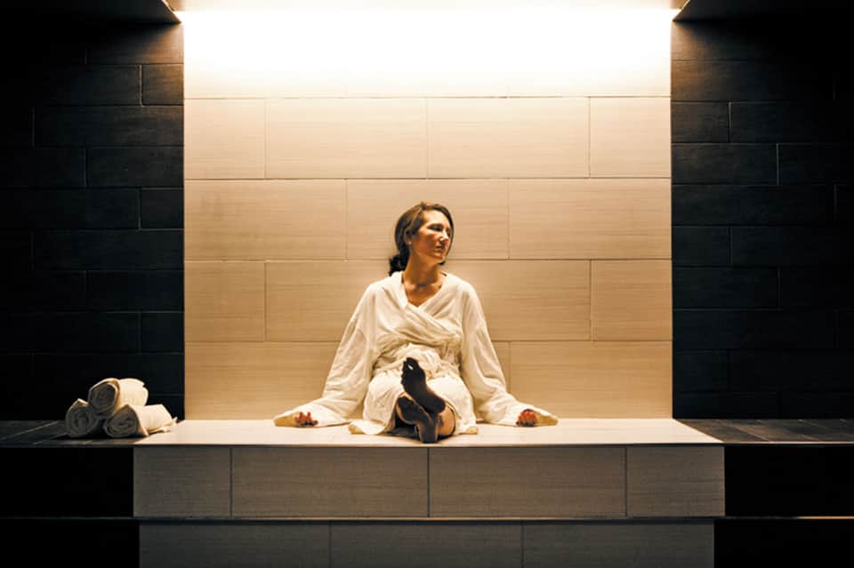Woman in white robe sitting on illuminated spa platform with rolled towels and black slippers