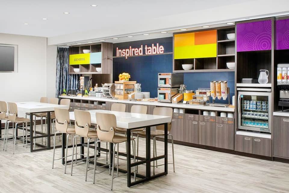 Modern hotel break room with colorful shelving, kitchenette, high-top dining table, and beige chairs