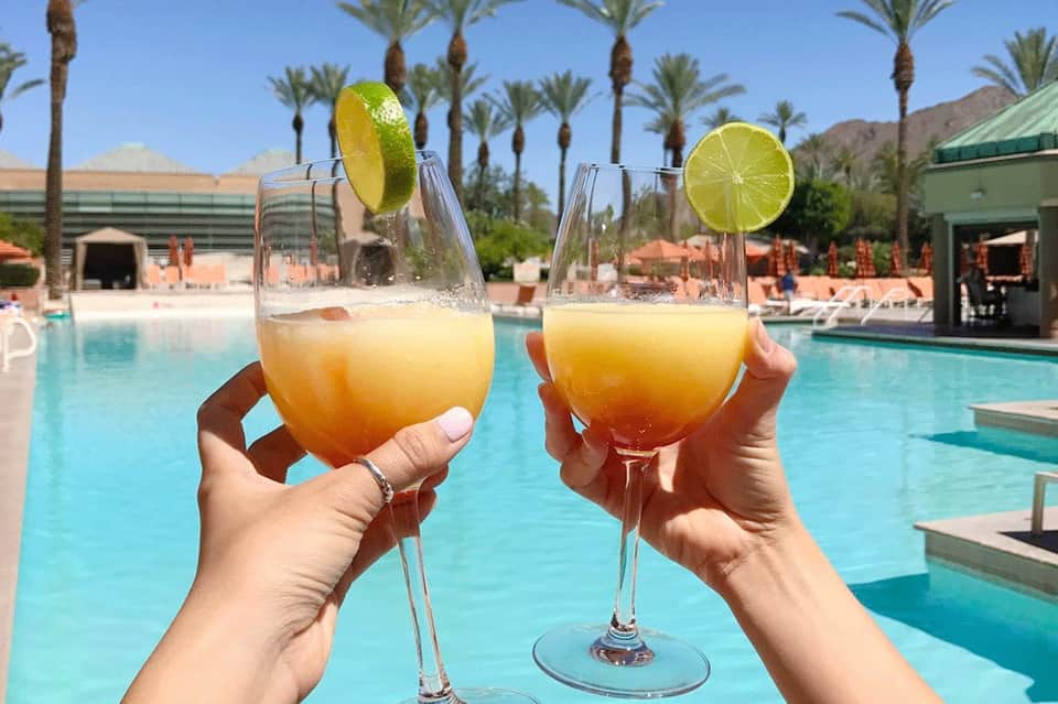 Two tropical cocktails with lime slices by a resort pool surrounded by palm trees and desert mountains