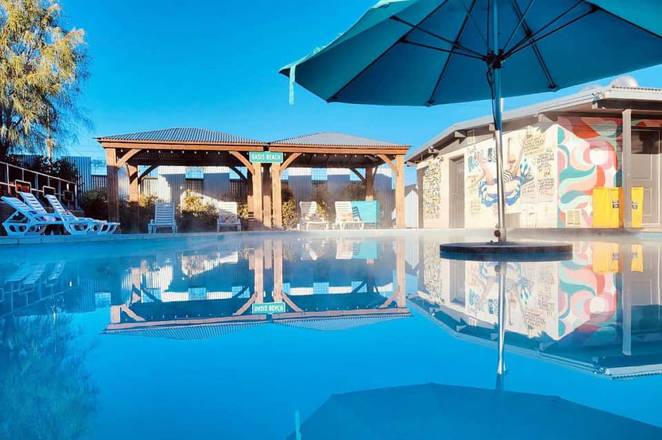 Modern pool with wooden pergola, blue umbrella, and reflected sky creating mirror-like surface