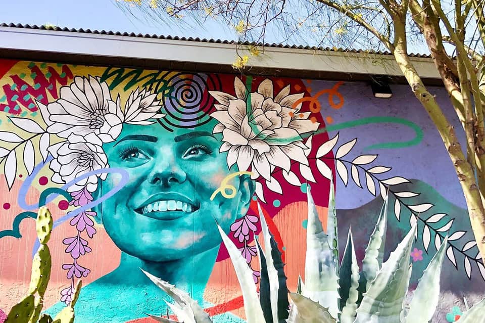 Vibrant street mural of turquoise smiling face with white flowers and colorful swirls