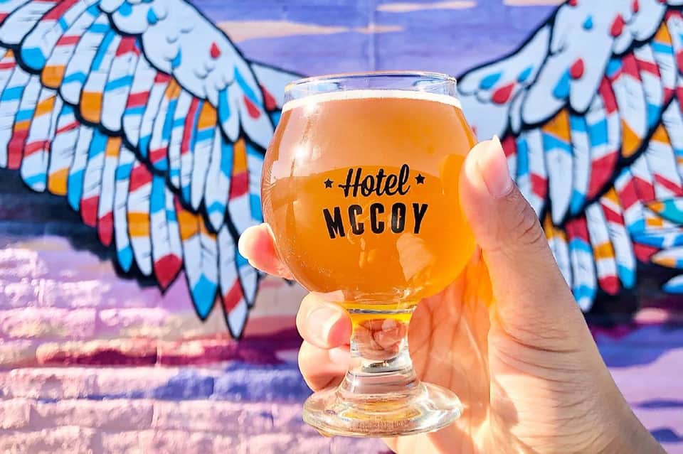 Beer glass labeled Hotel McCoy held up against colorful painted wings mural backdrop